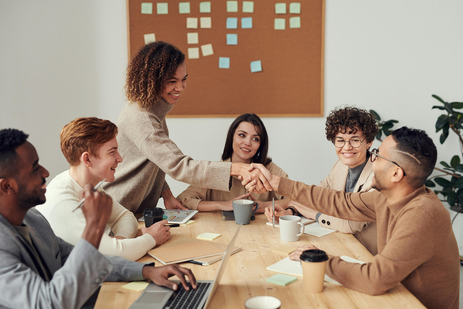 Business team meeting with diverse professionals shaking hands across conference table, cork board with sticky notes in background.