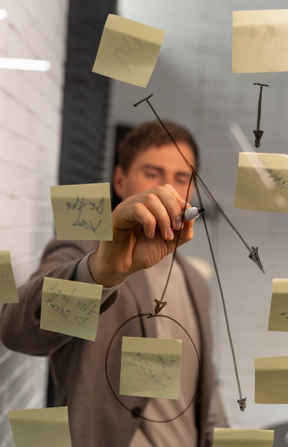 Person drawing connections between sticky notes on glass wall, holding marker while brainstorming or mapping ideas.